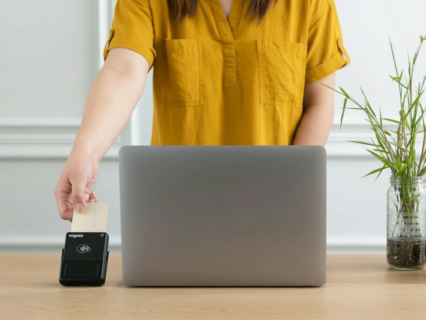 A person in a mustard yellow shirt swipes a card through a payment terminal next to a laptop on a wooden desk