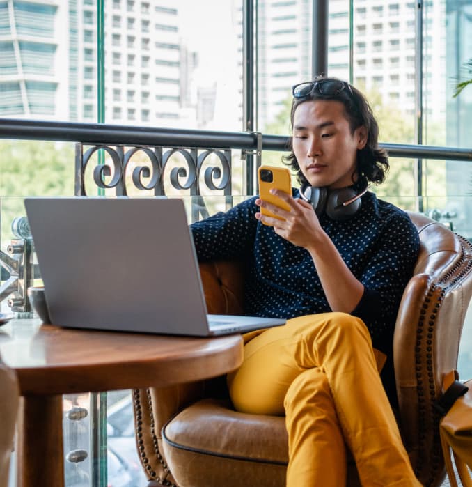 A person sits in a caf\u00e9 on a leather chair, holding a yellow phone and looking at it, with ai agents displayed on their laptop open on the wooden table. Headphones rest around their neck, city buildings visible through large windows.